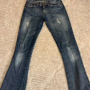 Blå bootcut jeans med slitningar - Snygga blå jeans med bootcut passform och slitna detaljer på benen. Jeansen har klassisk femficksdesign, låg midja och coolt broderi på bakfickan. Perfekta för dig som gillar en avslappnad och trendig look.
