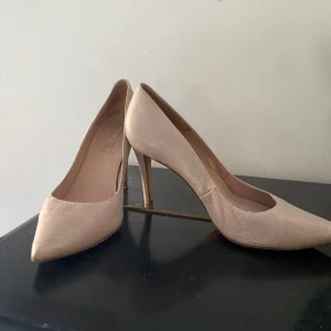 Beige pumps från Pura López - Snygga pumps i beige skinn från Pura López med spetsig tå och hög klack. Skorna har en stilren, vintage och klassisk design med lätt texturerad yta. Perfekta för dig som vill ha en elegant look med en neutral färg som passar till det mesta.