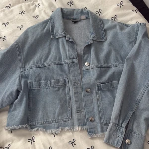 Ljusblå croppad jeansjacka - Säljer en ljusblå croppad jeansjacka med rå fransad nederkant och stora fickor framtill. Jackan har klassisk krage, knäppning med silvriga knappar och långa ärmar med manschett. Perfekt för dig som gillar en chill och trendig look.