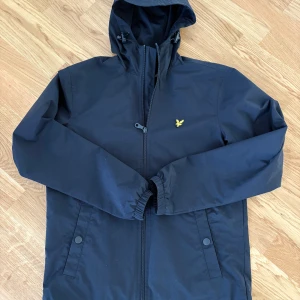 Mörkblå vindjacka från Lyle & Scott XS - Mörkblå vindjacka från Lyle & Scott med huva och dragkedja framtill. Jackan har två fickor med tryckknappar och den klassiska gula loggan på bröstet. Perfekt för blåsiga dagar och enkel att matcha med allt. Nyskick. 