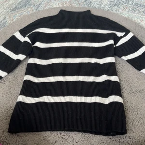 Svartvit randig polotopp från Vila - Säljer en svartvit polotopp från Vila med breda horisontella ränder. Toppen är ribbad och har lång ärm samt en hög krage. Materialet är stickad polyester som ger en skön och mjuk känsla. Perfekt för dig som gillar klassisk stil med en modern twist. har bara denna lapp men tror den är storlek S om inte större