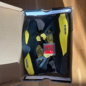 Nike Air Jordan 4 Retro 'Thunder' 45 - Säljer ett par Nike Air Jordan 4 Retro 'Thunder' i svart och gul. Skorna har meshpaneler, snörning och ikoniska Jumpman-loggor på hälen och innersulan. Ovandelen är i mocka och detaljerna i gult ger en riktigt fet kontrast. Perfekta för dig som vill sticka ut med din sneakerstil.