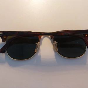 Ray-Ban Clubmaster solglasögon brun/guld - Snygga Ray-Ban Clubmaster solglasögon med klassisk brunsköldpaddsmönstrad båge och guldfärgade detaljer. Glasen är mörka och har en retroinspirerad form. Perfekt accessoar för en cool och tidlös look. 