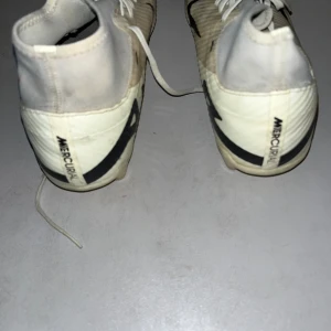 Nike Mercurial vita fotbollsskor - Säljer ett par Nike Mercurial fotbollsskor i vitt med svarta detaljer och snörning. Skorna har en högre strumpliknande krage och är tillverkade i syntetmaterial för en lätt känsla på planen. Perfekta för dig som vill sticka ut med stil på fotbollsplanen.