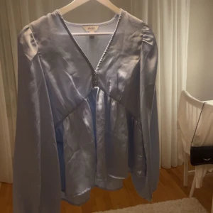 Ljusblå blus  - Supersöt ljusblå blus från AWARE med v-ringning och långa ärmar. Blusen har en glansig satinlik finish och volangdetalj nedtill som ger en flowy känsla. Perfekt att styla med jeans eller kjol för en trendig look. Knappt använd. 