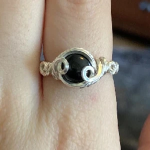 Silvrig ring med svart sten - Unik silvrig ring med en svart rund sten i mitten och snygga snirkliga detaljer på sidorna. Ringen har en bohemisk och handgjord känsla som sticker ut och passar perfekt för dig som gillar personliga accessoarer.