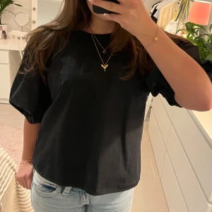 Svart blus med puffärm - Svart blus med puffiga korta ärmar och rund halsringning. Baksidan har en snygg öppning med knytband i nacken. Blusen är i ett lätt bomullstyg och har en avslappnad passform som ger en cool och trendig vibe. 