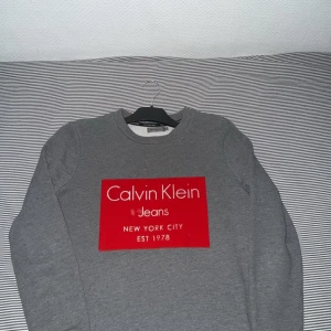 Grå Calvin Klein Jeans sweatshirt - Snygg grå sweatshirt från Calvin Klein Jeans med stort rött tryck framtill. Tröjan har rund halsringning och långa ärmar. Perfekt för dig som gillar stilrena och klassiska plagg med en modern touch.