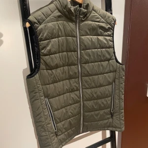 Olivgrön quiltad dunväst från Zara - Olivgrön quiltad dunväst från Zara med hög krage och dragkedja framtill. Västen har två fickor med dragkedja och en stilren, minimalistisk look. Perfekt att slänga över en hoodie eller tröja för extra värme.