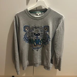 Grå Kenzo sweatshirt med tigertryck - Säljer en grå sweatshirt från Kenzo med ikoniskt broderat tigerhuvud i blått, grönt och gult framtill. Tröjan har rund halsringning, ribbade muddar och är långärmad. Perfekt statement-piece för dig som gillar streetstyle.