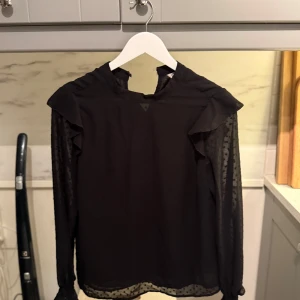 Svart blus med volang och prickiga ärmar - Snygg svart blus med volangdetaljer på axlarna och långärmade ärmar med diskret prickigt mönster, från Cubus. Passar perfekt till kalas och fester!