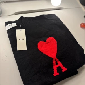 Svart stickad tröja med rött hjärta från Ami - Svart stickad tröja från Ami Alexandre Mattiussi med ett stort rött hjärta och bokstaven A på bröstet. Tröjan är tillverkad i ekologisk bomull och har ribbade muddar vid hals, ärmslut och nederkant. Perfekt statement-piece för dig som gillar stilrena detaljer.