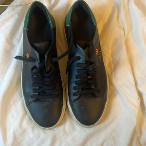 Svarta Axel Arigato sneakers med broderad blomma - Svarta sneakers från Axel Arigato med vit sula och gröna detaljer vid hälen. Sidan pryds av en broderad glad blomma i rött, gult och grönt. Skorna har svarta skosnören och är tillverkade i skinn. Perfekta för dig som vill ha en unik och lekfull touch på dina sneakers.