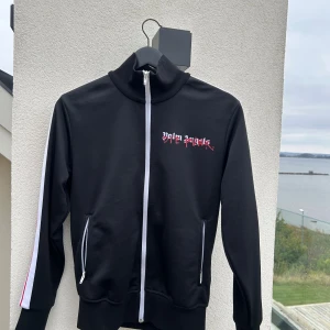 Palm Angels jacka  - Svart track jacket från Palm Angels med vit dragkedja, vita och röda ränder längs ärmarna och logga på bröstet. Hög krage och två fickor med dragkedja. Tillverkad i 100% polyester, perfekt för en sportig och trendig look. Collab med Playboy Carti. Köpt i New York, köpt för 12 000kr.