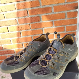 Grå mesh-sneakers från ECCO eUR 42 - Säljer ett par grå sneakers från ECCO med meshpaneler och snygga detaljer i beige och gulbrunt. Skorna har snörning med elastiska snören och en platt, slitstark sula. Perfekta för dig som gillar sportig och bekväm stil. ECCO-logga på både plös och häl.