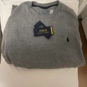 Grå sweatshirt från Polo Ralph Lauren - Snygg grå sweatshirt från Polo Ralph Lauren med klassisk rund hals och broderad marinblå logga på bröstet. Tröjan har ribbade muddar vid ärmslut och nederkant. Perfekt för en chill och stilren look.