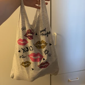 Grå handgjord tygkasse - Säljer en upcycled grå tygkasse i mjuk bomull med tryck av rosa och leopardmönstrade läppar, små hjärtan och text som 'XOXO''. Perfekt för att bära med sig allt man behöver och samtidigt bra för miljön, då den är handsydd från en T-shirt. 