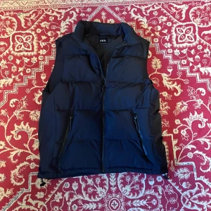 Svart dunväst från Zara - Snygg svart dunväst från Zara med hög krage och två dragkedjefickor framtill. Västen är quiltad och har en stilren look med rak passform. Perfekt att slänga över en hoodie eller tröja när det är kyligt ute.