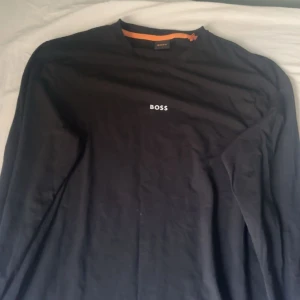 Svart långärmad tröja från BOSS - Svart långärmad tröja från BOSS med enkel och stilren design. Tröjan har rund halsringning och en liten vit BOSS-logga på bröstet. Perfekt för dig som gillar klassiska och cleana plagg med diskret branding.