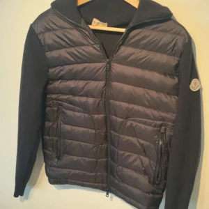 Svart cardigan från Moncler med huva - Svart dunjacka från Moncler med stickade ärmar och huva. Jackan har quiltad framsida, två fickor med dragkedja och Moncler-logga på ärmen. Perfekt för dig som vill ha en snygg och varm jacka med street vibe och exklusiv känsla.