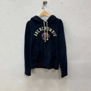 Abercrombie & Fitch Hoodie - Mörkblå hoodie från Abercrombie & Fitch med broderad logga och emblem framtill | Storlek M|  Sliten still på tröjan| Hör av dig vid frågor eller funderingar 🤩!