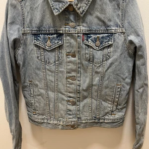 Klassisk jeansjacka från Levi's - Säljer en klassisk ljusblå jeansjacka från Levi's med snygga bröstfickor och knappar framtill. Jackan har en tidlös design med krage och raka sömmar, perfekt för dig som gillar streetstyle och denim. Ikonisk röd Levi's-tagg på bröstfickan. Aldrig använd