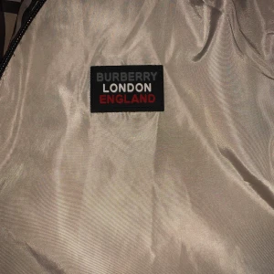 Beige Burberry vindjacka med huva - Snygg beige vindjacka från Burberry med klassiskt rutigt foder i huvan och svarta dragkedjor. Jackan har en patch med Burberry London England på bröstet och två fickor framtill. Perfekt för dig som vill sticka ut med stilren design.