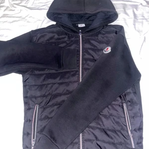 Svart hoodiejacka från Moncler - Säljer en svart hoodiejacka från Moncler med quiltad framsida och mjuka ärmar. Jackan har dragkedja framtill och två fickor med dragkedja. Klassisk Moncler-logga på bröstet och huva för extra stilpoäng.