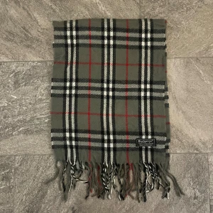 Rutig halsduk från Burberrys i ull - Klassisk rutig halsduk från Burberrys i mjuk ull. Färgerna är grå, svart, vit och röd med fransar i ändarna. Perfekt accessoar för att lyfta din outfit med en tidlös brittisk vibe. Tydlig Burberrys-logga på etiketten.