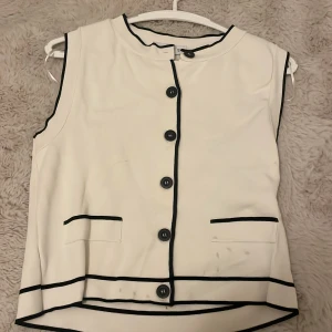 Vit tröjamed svarta detaljer från MNG - Vit tröja  från MNG med svarta kontrastkanter och svarta knappar framtill. Tröjan har två fickor framtill och är ärmlös med rund hals. Snyggt grafiskt uttryck med raka linjer och klassisk passform. Fläckarna går att ta bort med nål specifik medel. Kan göra det om man är intresserad och vill köpa!!🥰 Använt endast en gång