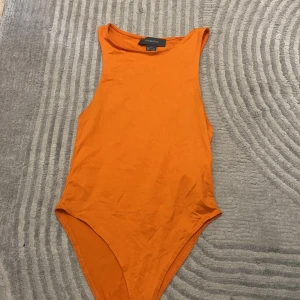Orange ärmlös bodysuit från Primark - Säljer en snygg orange bodysuit från Primark i storlek 2XS. Modellen är ärmlös med rund hals och har praktiska tryckknappar i grenen. Materialet är mjukt och stretchigt, perfekt för att styla med jeans eller kjol. Färgen är riktigt poppande orange och passformen är tight.