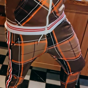 The Gentlemen Tracksuit/Jogging Dress/Two piece [Medium]  - Tracksuit med rutigt mönster i orange, svart och vitt inspirerad av filmen The Gentlemen. Dressen har vita och röda ränder längs sidorna och dragsko i midjan. Sköna och snygga för dig som gillar sportig stil och vill sticka ut. Utmärkt skick. 