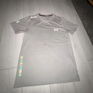 Grå t-shirt från Under Armour - Snygg grå t-shirt från Under Armour med korta ärmar och rund hals. Logga på bröstet och färgglad text längs sidan. Perfekt för dig som gillar sportig och clean stil.