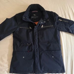 Snoot Vinterjacka - Snygg marinblå vinterjacka från Snoot med orange quiltat foder. Jackan har flera dragkedjefickor, hög krage och coola metallknappar. Materialet är polyester och jackan har en modern, normal passform. Perfekt för dig som gillar stilren och funktionell design.