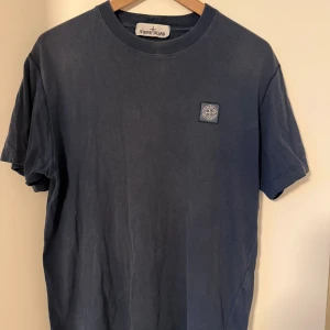 Blå Stone Island t-shirt i bomull - Säljer en marinblå t-shirt från Stone Island med klassisk logopatch på bröstet. T-shirten är i mjuk bomull och har rund hals och korta ärmar. Perfekt för dig som gillar streetwear och vill ha en clean look med ikonisk branding.