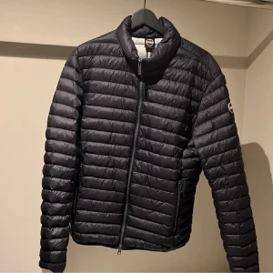 Svart dunjacka från Colmar, stl 50 - Snygg svart dunjacka från Colmar Originals med horisontella sömmar och diskret logga på ärmen. Jackan har dragkedja framtill, två sidofickor med dragkedja och stårkrage. Lätt och smidig modell som passar perfekt till kyliga dagar.