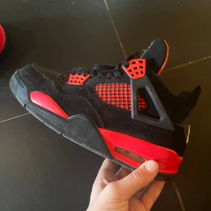 Nike Air Jordan 4 svart/röd sneakers - Säljer ett par Nike Air Jordan 4 sneakers i svart och rött med klassisk Jumpman-logga på plösen och hälen. Skorna har meshpaneler, snörning och detaljer i rött samt svart mocka och syntetmaterial. Perfekta för dig som vill sticka ut med en ikonisk streetwear-look.