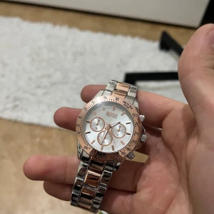 Hugo Boss klocka i silver och roséguld - Snygg klocka från Hugo Boss med länkarmband i silver och roséguld. Urtavlan är vit med roséguldsdetaljer och tre mindre urtavlor. Boetten är rund och har flera knappar på sidan. Perfekt accessoar för dig som gillar stilrena detaljer.