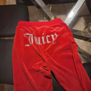 Röda Juicy Couture mjukisbyxor S - Röda mjukisbyxor från Juicy Couture i sammet med glittrande Juicy-text baktill och strassdetalj fram. Byxorna har snörning i midjan och fickor på sidorna. Perfekta för en avslappnad och trendig look.