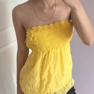 Gul stickad bandeau tubtopp med blomma - Säljer en söt gul bandeau tubtopp med stickad överdel och volangig, luftig nederdel. Toppen har en dekorativ stickad blomma framtill och vågig kant upptill. Perfekt för sommardagar och enkel att styla med jeans eller kjol.