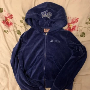 Blå velour hoodie Juicy Couture - Mörkblå hoodie i mjuk velour från Juicy Couture med glittrande stenar och broderad krona på huvan. Dragkedja framtill, fickor och snyggt dekorerad rygg med logga och stenar. Perfekt för dig som vill ha en ikonisk och lyxig streetstyle-look.