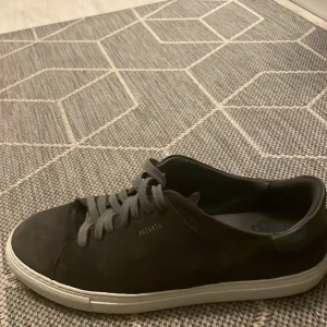 Svarta sneakers från Arigato - Snygga svarta sneakers från Arigato med stilren design och vit platt sula. Skorna har rund tå, snörning och är tillverkade i mocka med detaljer i skinn. Perfekta för dig som gillar minimalistisk och modern stil.