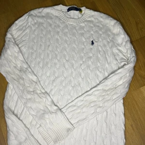 Vit tröja Polo Ralph Lauren - Vit kabelstickad tröja från Polo Ralph Lauren med klassisk rund halsringning och broderad marinblå logga på bröstet. Tröjan är i bomull och har långa ärmar samt ribbade muddar och nederkant. Perfekt för en clean och stilren look. Pris kan diskuteras fraktar samma dag 
