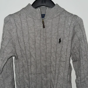 Grå kabelstickad tröja Polo Ralph Lauren - Kabelstickad grå tröja från Polo Ralph Lauren med halv dragkedja och hög krage. (Obs säljer pga för liten storlek, vid snabb affär kan priset definitivt diskuteras). Använd endast en gång.