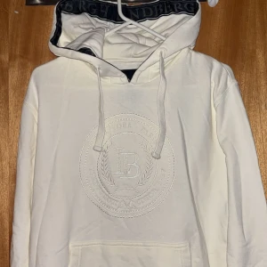 Vit hoodie från Lindbergh med broderi - Snygg vit hoodie från Lindbergh i storlek S med stort broderat emblem på bröstet. Tröjan har huva med snörning och ribbade muddar. Materialet är mjuk bomull och insidan av huvan har mörkblå detaljer med Lindbergh-tryck. Perfekt för chill och streetwear.