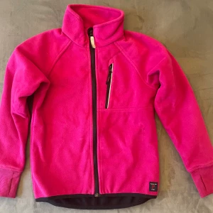 Rosa fleecejacka Polarn O. Pyret 128 - Mjuk och varm rosa fleecejacka från Polarn O. Pyret i storlek 128. Jackan har hel dragkedja framtill, hög krage och en bröstficka med dragkedja. Perfekt för kyliga dagar och enkel att matcha med andra plagg.