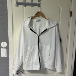 Vit hoodie från Stone Island - Vit hoodie från Stone Island med svart dragkedja och klassisk logga på vänster ärm. Tröjan har huva, två stora fickor framtill och är tillverkad i mjuk bomull. Perfekt för en clean och stilren look. Nytt skick, använd en gång.