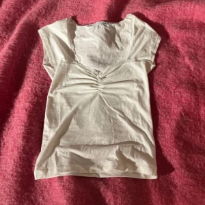 Vit topp med spets från 157 - Supersöt vit topp från 157 i storlek XS med v-ringning, spetskant och liten rosett framtill. Toppen har korta ärmar och är gjord i mjuk bomull. Rynkade detaljer vid bysten ger en extra touch. Perfekt för dig som gillar en feminin och enkel stil.