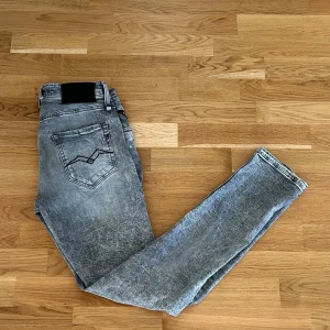 Grå slim jeans från replay - Grå jeans från replay i slim tapered fit med slitna detaljer och tvättad look. Klassisk femficksmodell med snygga sömmar på bakfickan. Strl: 29 