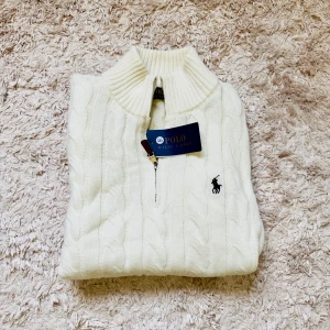 Vit stickad polotröja Polo Ralph Lauren - Säljer en vit stickad polotröja från Polo Ralph Lauren med klassiskt kabelstickat mönster, halv dragkedja med läderdetalj och broderad marinblå logga på bröstet. Perfekt för dig som vill ha en clean och tidlös look.